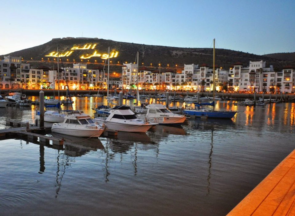 View of Agadir Marina with sailboats docked along floating piers, surrounded by white Mediterranean-style apartment buildings with red roofs, set against a rocky hillside and a bright blue sky.